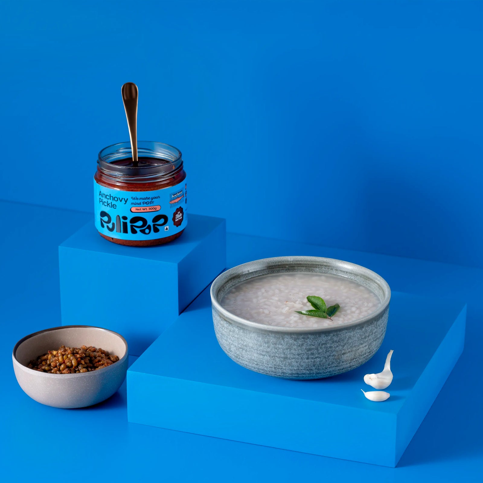 Anchovy Pickle by PuliPOP with a bowl of rice kanji and lentils on blue stepped pedestals